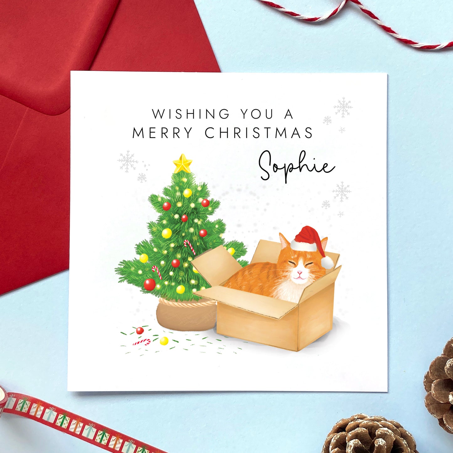 A personalised Christmas card featuring a ginger and white tabby cat in a box wearing a santa hat next to a christmas tree. It can be personalised with a name, and is suitable for a range of relations such as Wife, Girlfriend, Daughter, Granddaughter, Niece and Mum.