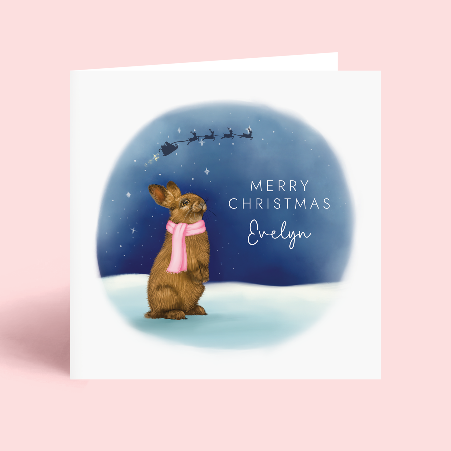 A Personalised Cute Christmas Card, featuring a detailed illustration of a brown rabbit wearing a pink scarf, looking up at a starry sky. The silhouette of Santa's sleigh can be seen in the sky. It can be personalised with any girl's name.