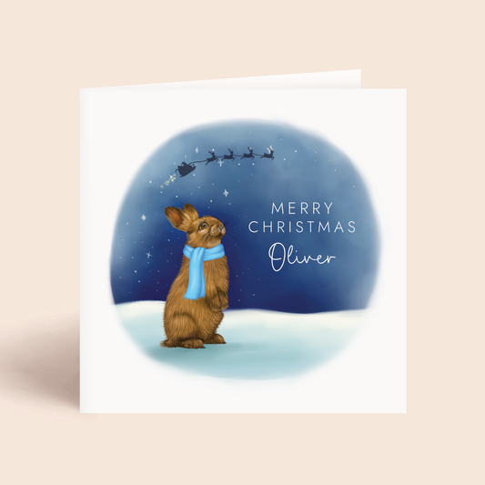 Personalised Christmas Card, featuring a detailed illustration of a brown rabbit wearing a blue scarf, looking up at a starry sky. The silhouette of Santa's sleigh can be seen in the sky. It can be personalised with any boy's name.
