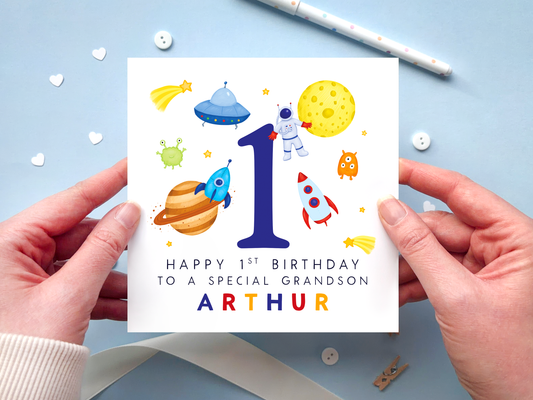 A bold and bright space themed 1st birthday card for boys. It features an astronaut, moon, planet, aliens, rockets and stars, around the number one. It can be personalised with the child's name.