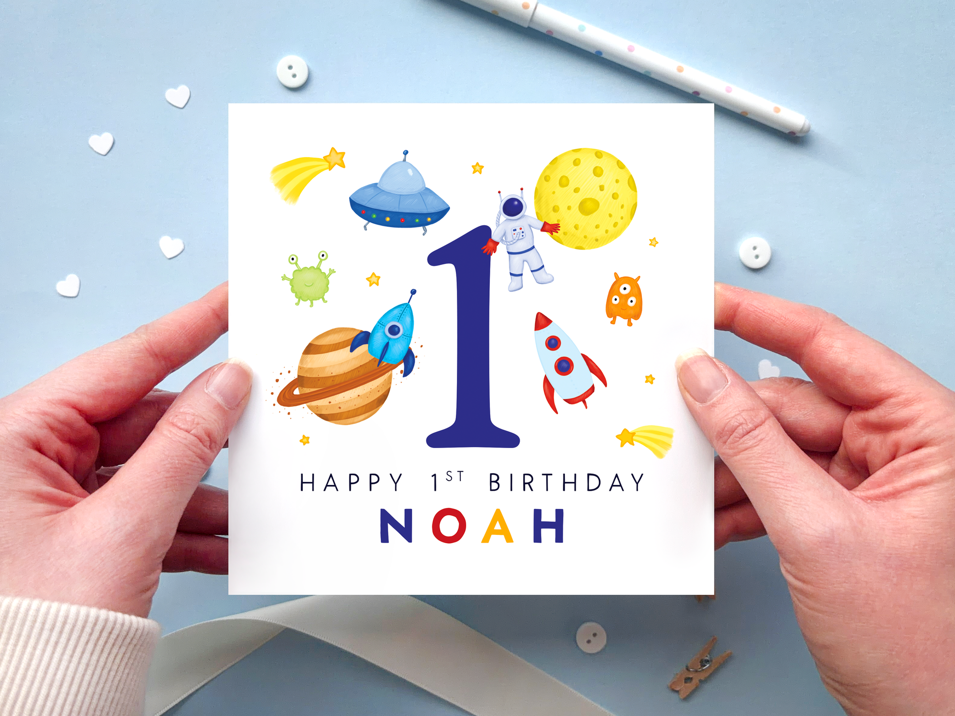 A bold and bright space themed 1st birthday card for boys. It features an astronaut, moon, planet, aliens, rockets and stars, around the number one. It can be personalised with the child's name.