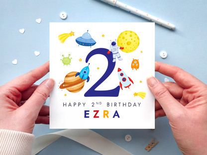 A bold and bright space themed 2nd birthday card for boys. It features an astronaut, moon, planet, aliens, rockets and stars, around the number two. It can be personalised with the child's name.
