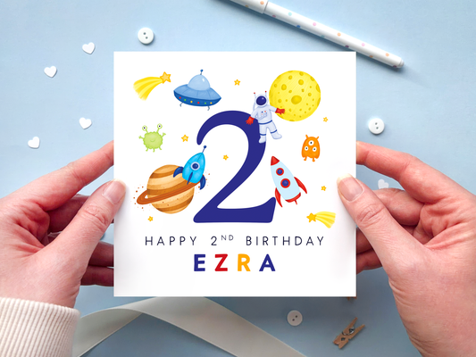 A bold and bright space themed 2nd birthday card for boys. It features an astronaut, moon, planet, aliens, rockets and stars, around the number two. It can be personalised with the child's name.