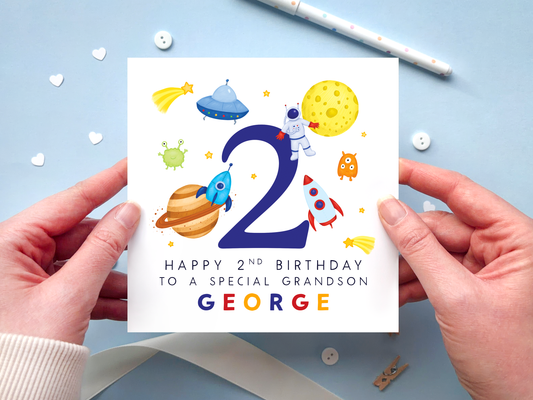 A bold and bright space themed 2nd birthday card for boys. It features an astronaut, moon, planet, aliens, rockets and stars, around the number two. It can be personalised with the child's name.