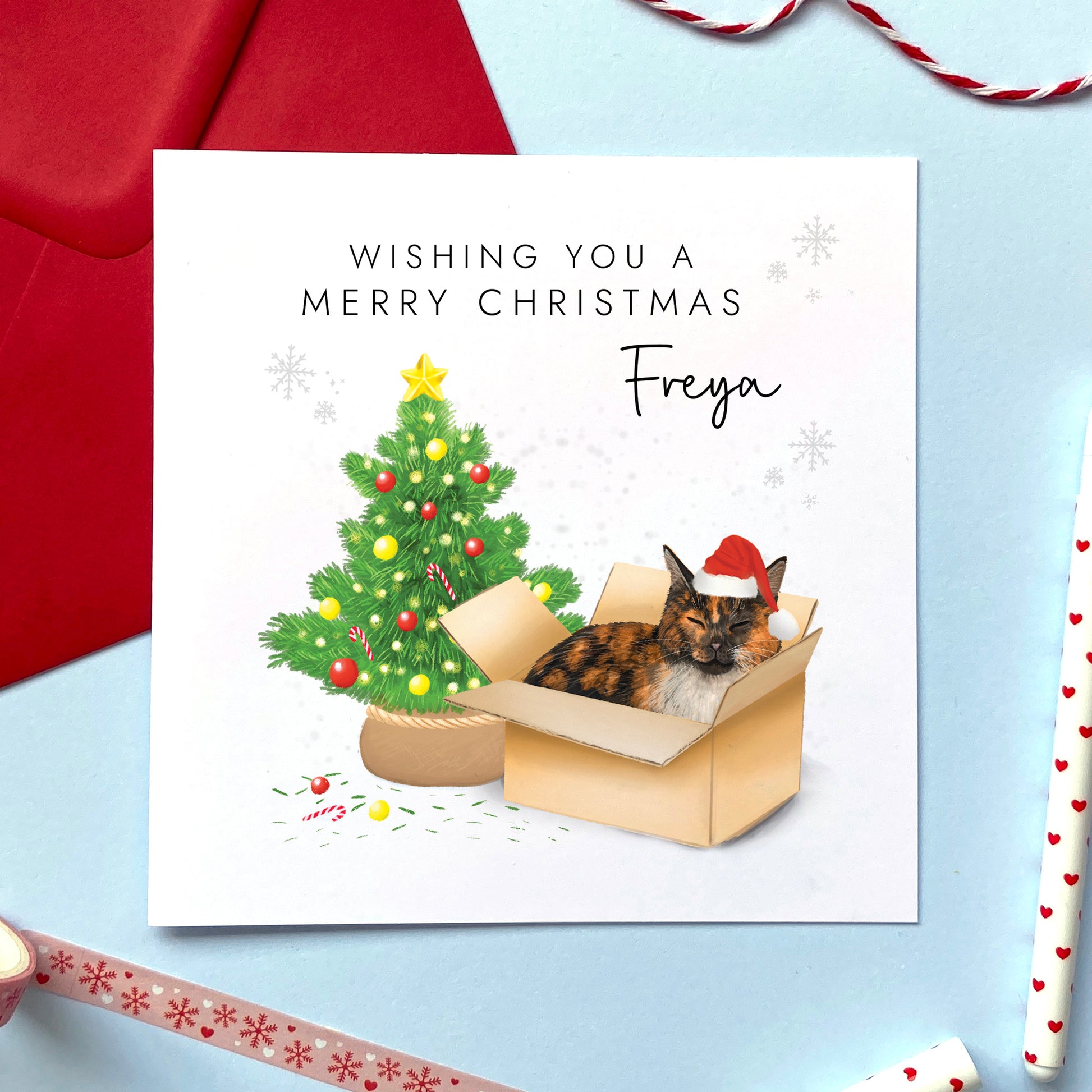 A personalised Christmas card featuring a black, white and ginger tortoiseshell cat in a box wearing a santa hat next to a christmas tree. It can be personalised with a name, and is suitable for a range of relations such as Wife, Girlfriend, Daughter, Granddaughter, Niece and Mum.